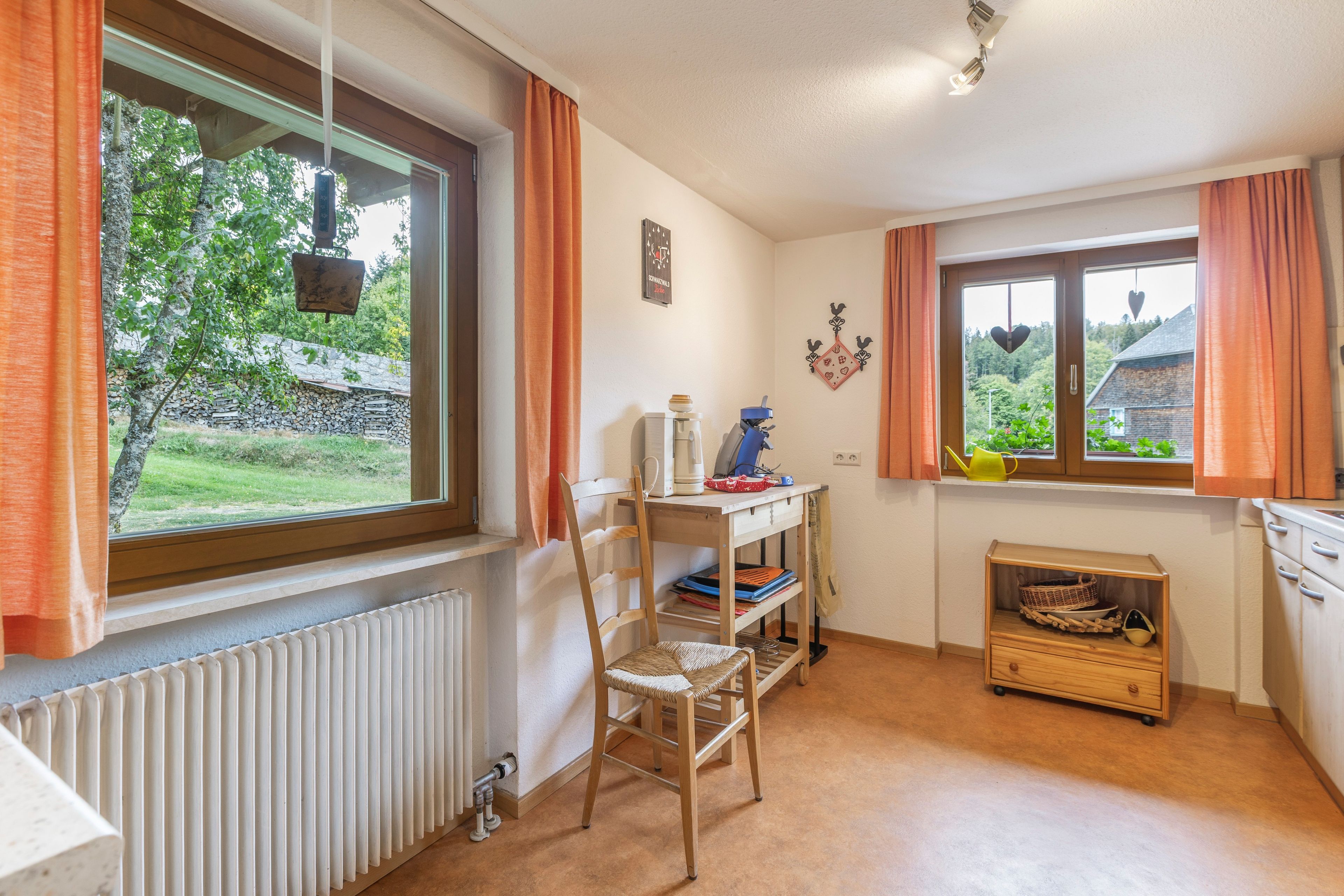 Ein Zimmer mit Heizkörper, einem Holzstuhl, einem Tisch mit Wasserkocher und einem Fenster mit Vorhängen.
