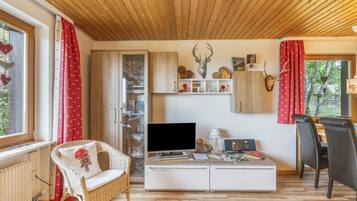 Una sala de estar acogedora con televisor, un mueble blanco, un sillón de mimbre y un comedor con mesa y sillas.