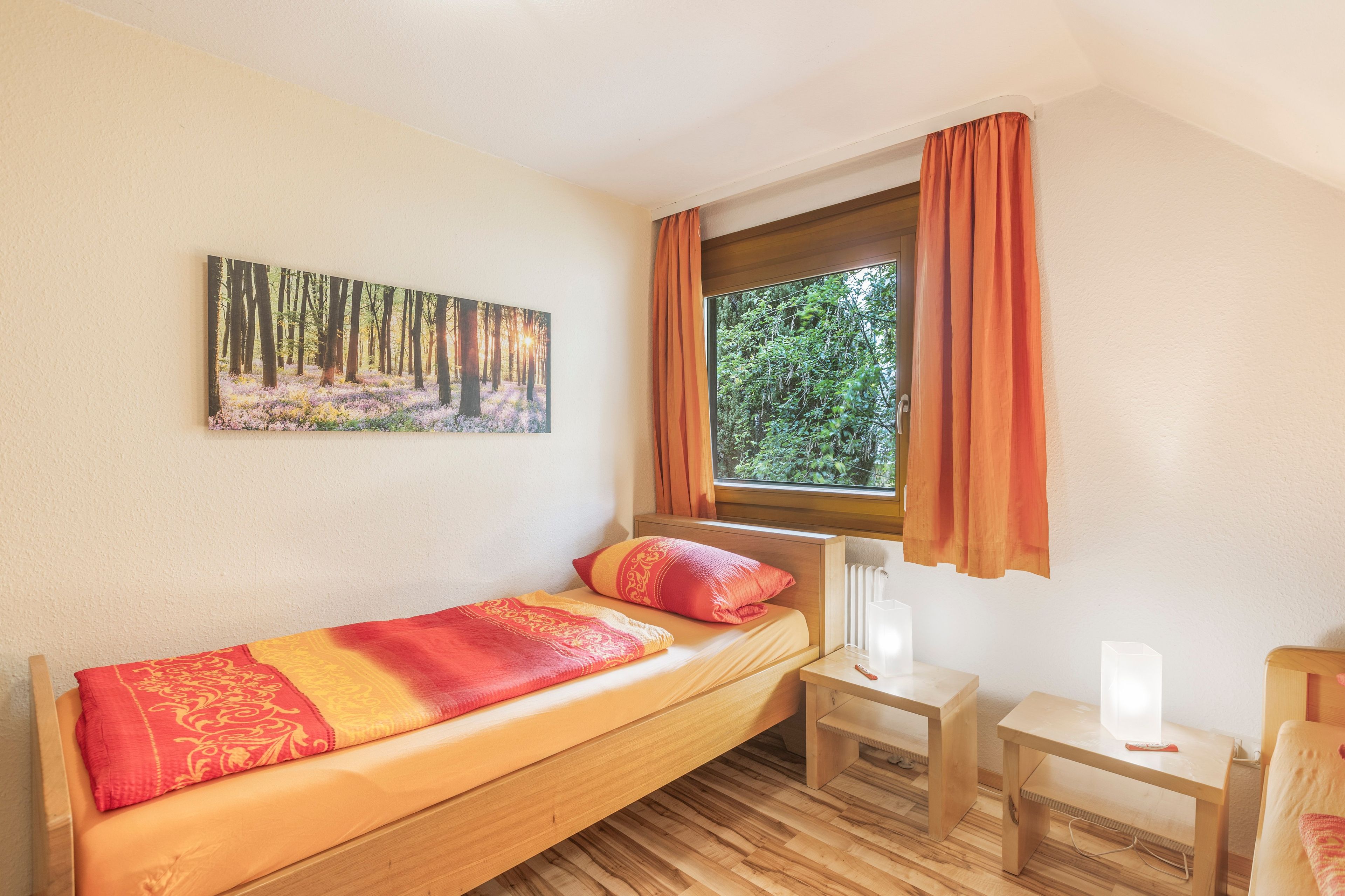 Ein Einzelbett mit einem gemusterten Bettbezug, ein kleiner Nachttisch, ein Stuhl und ein Fenster mit Vorhängen.