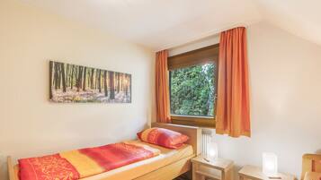 Ein Einzelbett mit einem gemusterten Bettbezug, ein kleiner Nachttisch, ein Stuhl und ein Fenster mit Vorhängen.