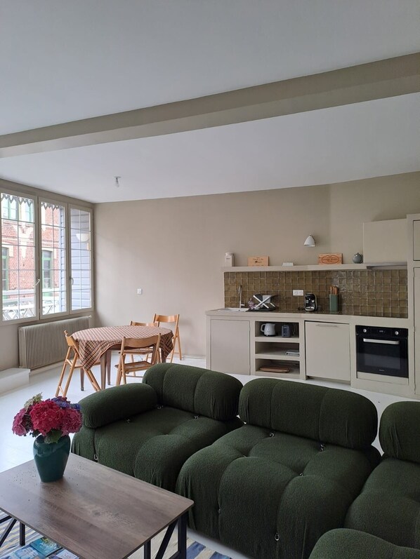 A modern living room with a green sofa set, a dining table with chairs, and a kitchen area with a tiled backsplash and appliances.