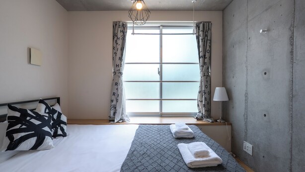 A hotel room with a bed, a window with curtains, a lamp, and a wall with a power outlet.