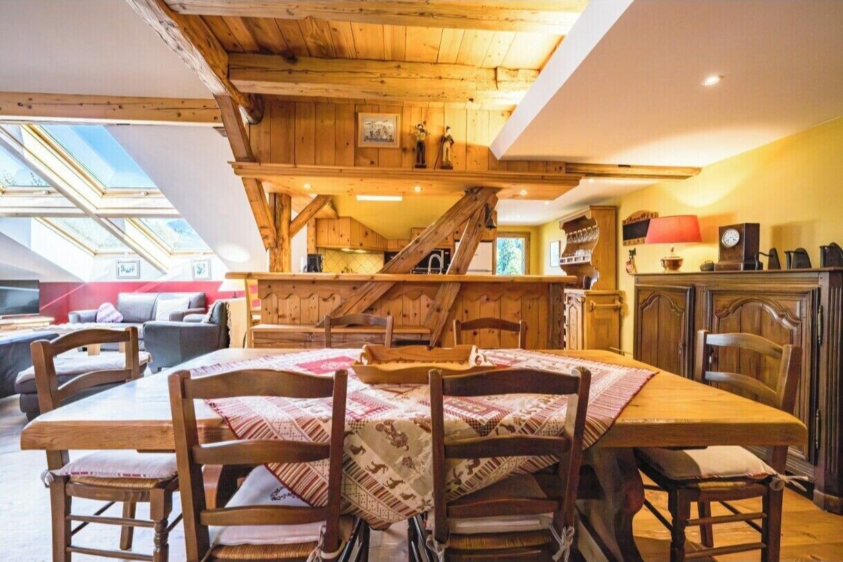 A cozy, rustic dining area with a wooden table and chairs, a kitchen in the background, and a skylight above.