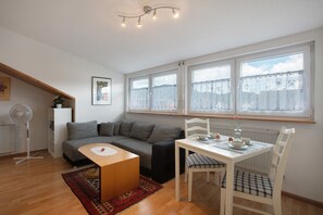 Un espace de vie compact comprenant un canapé, une table à manger et une chaise. On y trouve également un radiateur, un ventilateur et une fenêtre avec des rideaux.