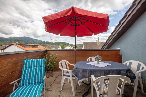 A balcony with a red umbrella, white chairs, a table with a blue tablecloth, and a potted plant.