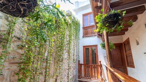 Un patio estrecho con un muro de piedra cubierto de plantas trepadoras, balcones de madera con barandillas y macetas colgantes.