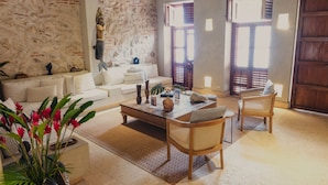 Una sala de estar con sofá blanco, una mesa de centro de madera, sillas de mimbre, una pared de piedra y una puerta de madera.