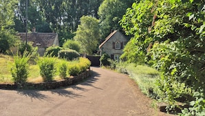 Un chemin de campagne mène à une maison en pierre, entourée d’une végétation luxuriante et d’arbres.