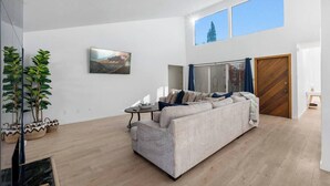 A spacious living room with a large sofa, a coffee table, and a painting on the wall. There are two windows providing natural light and a view of a building outside.
