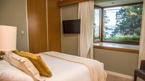 Quarto de hotel com uma cama, televisão na parede, janela com vista para vegetação e uma cadeira.