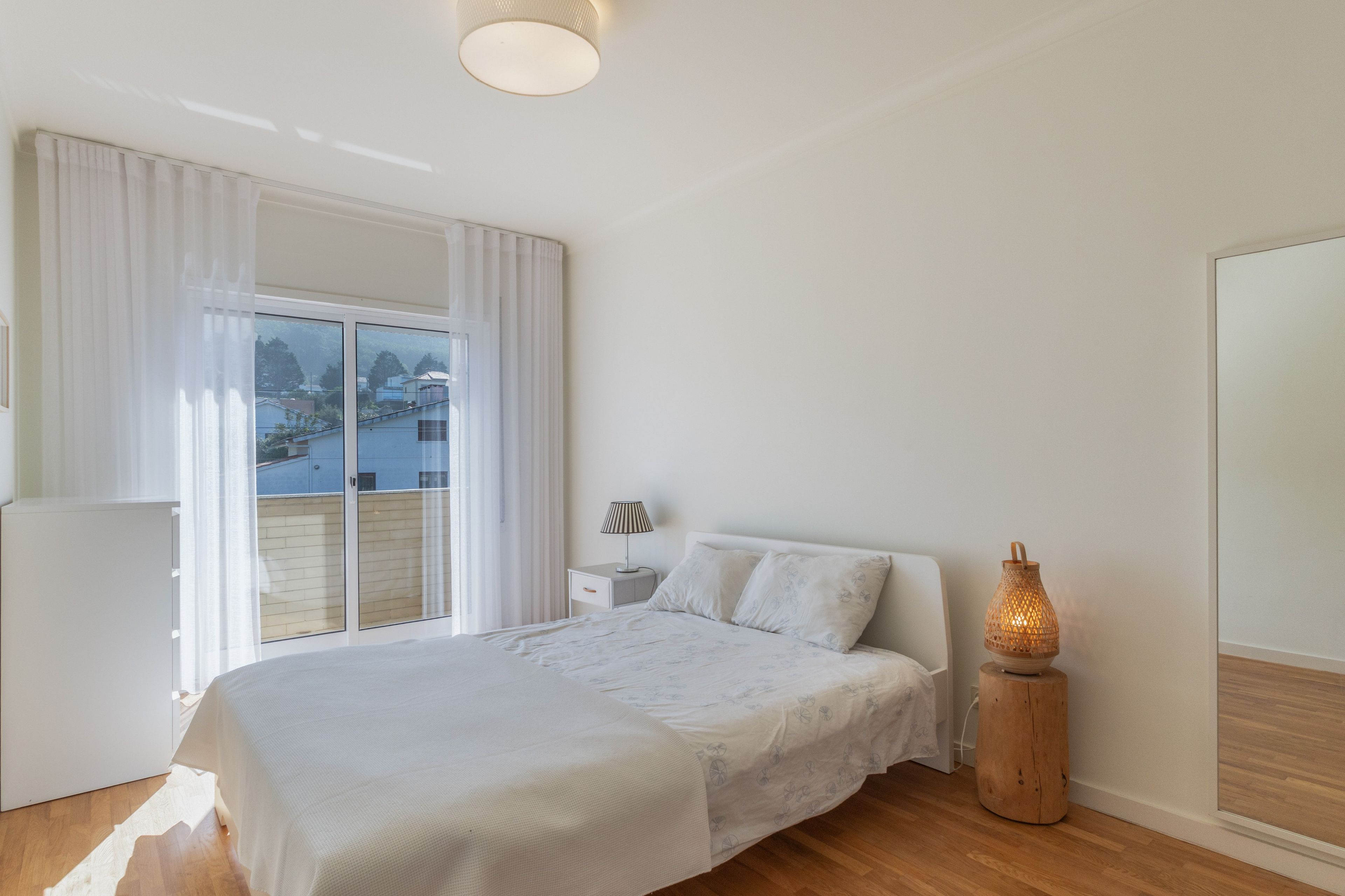 A bedroom with a bed, a nightstand, a lamp, and a view of the outside.