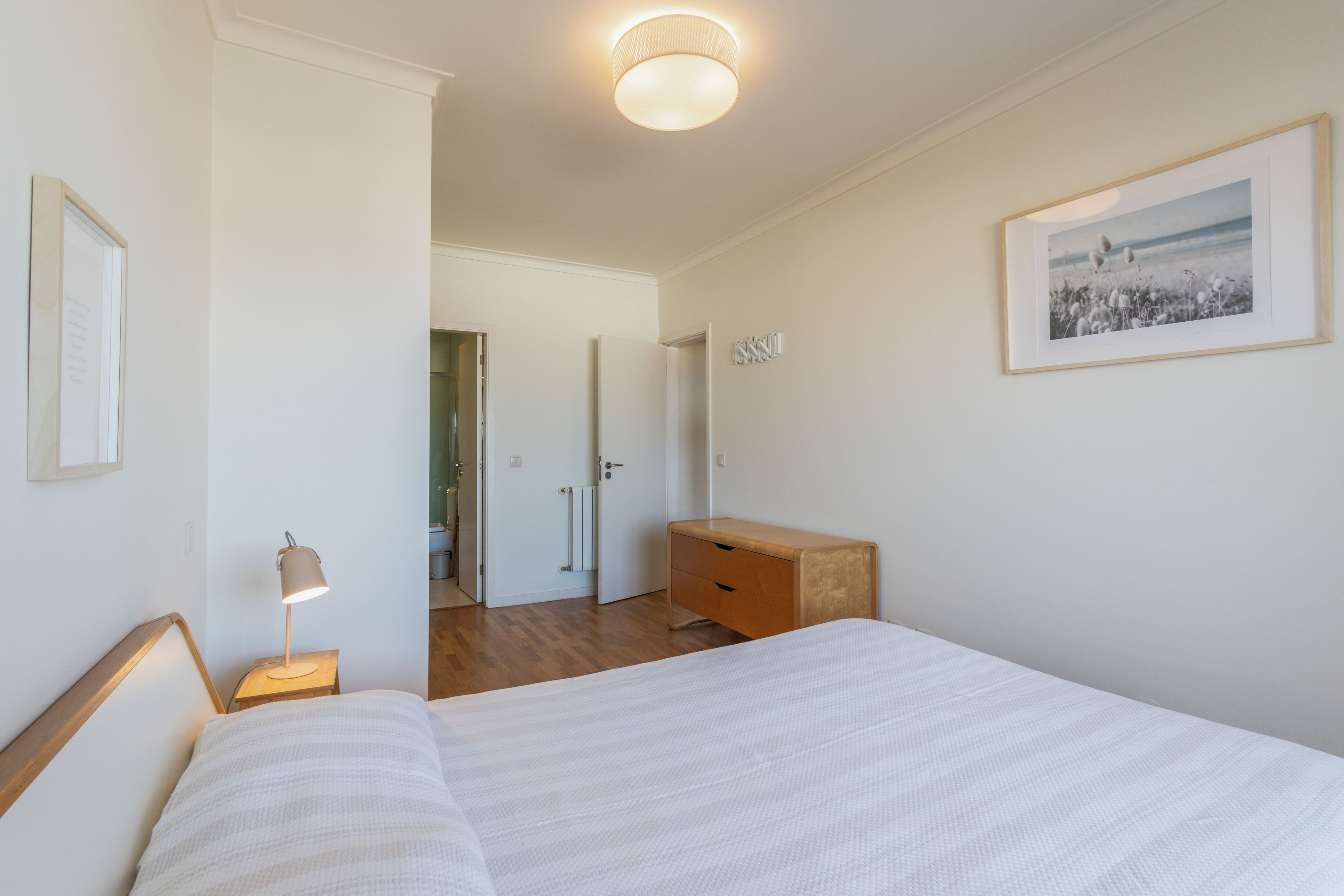 Un dormitorio con cama, mesita de noche, lámpara, cómoda y un cuadro en la pared.
