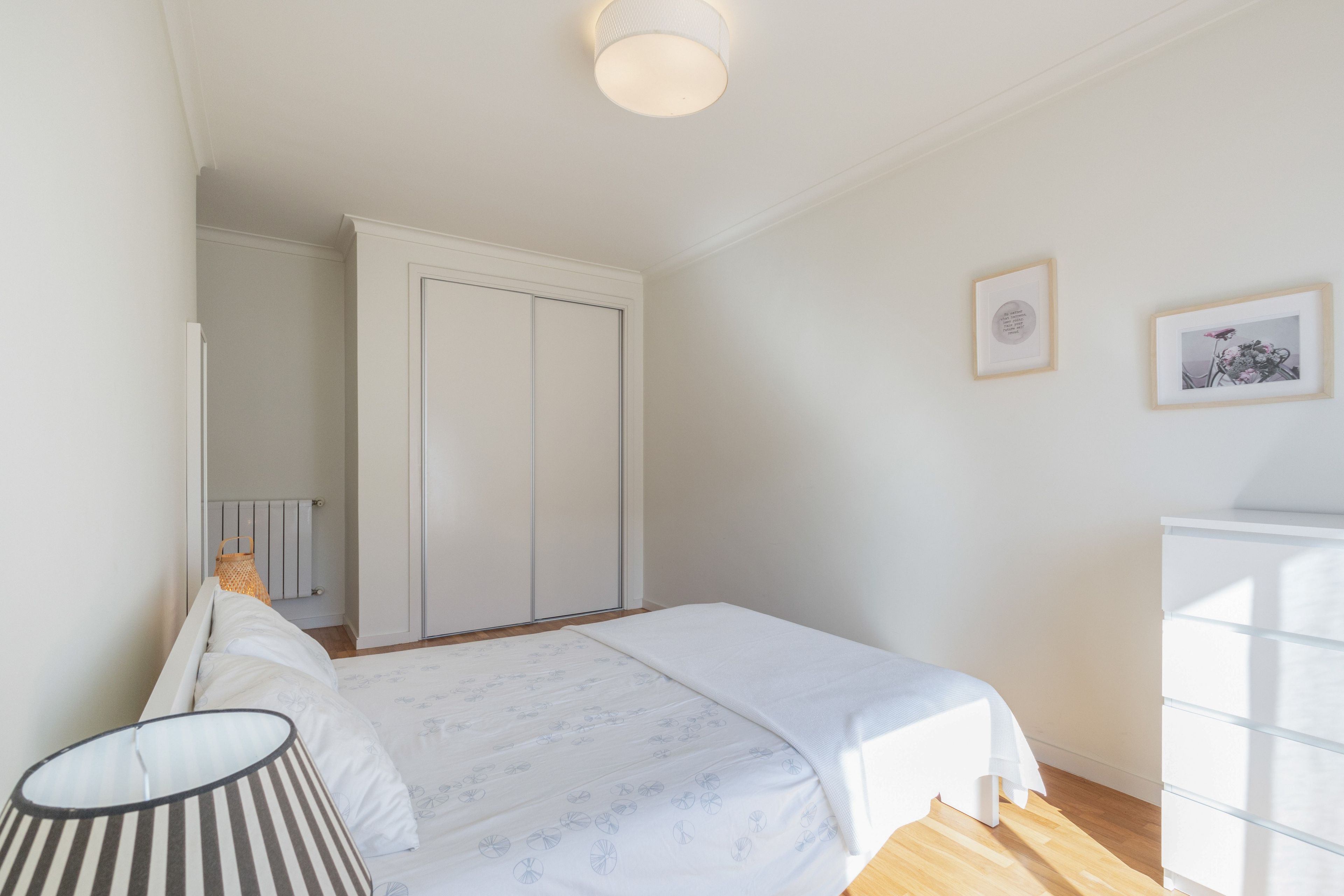 A bedroom with a bed, a bedside lamp, a radiator, a wardrobe, and two framed pictures on the wall.