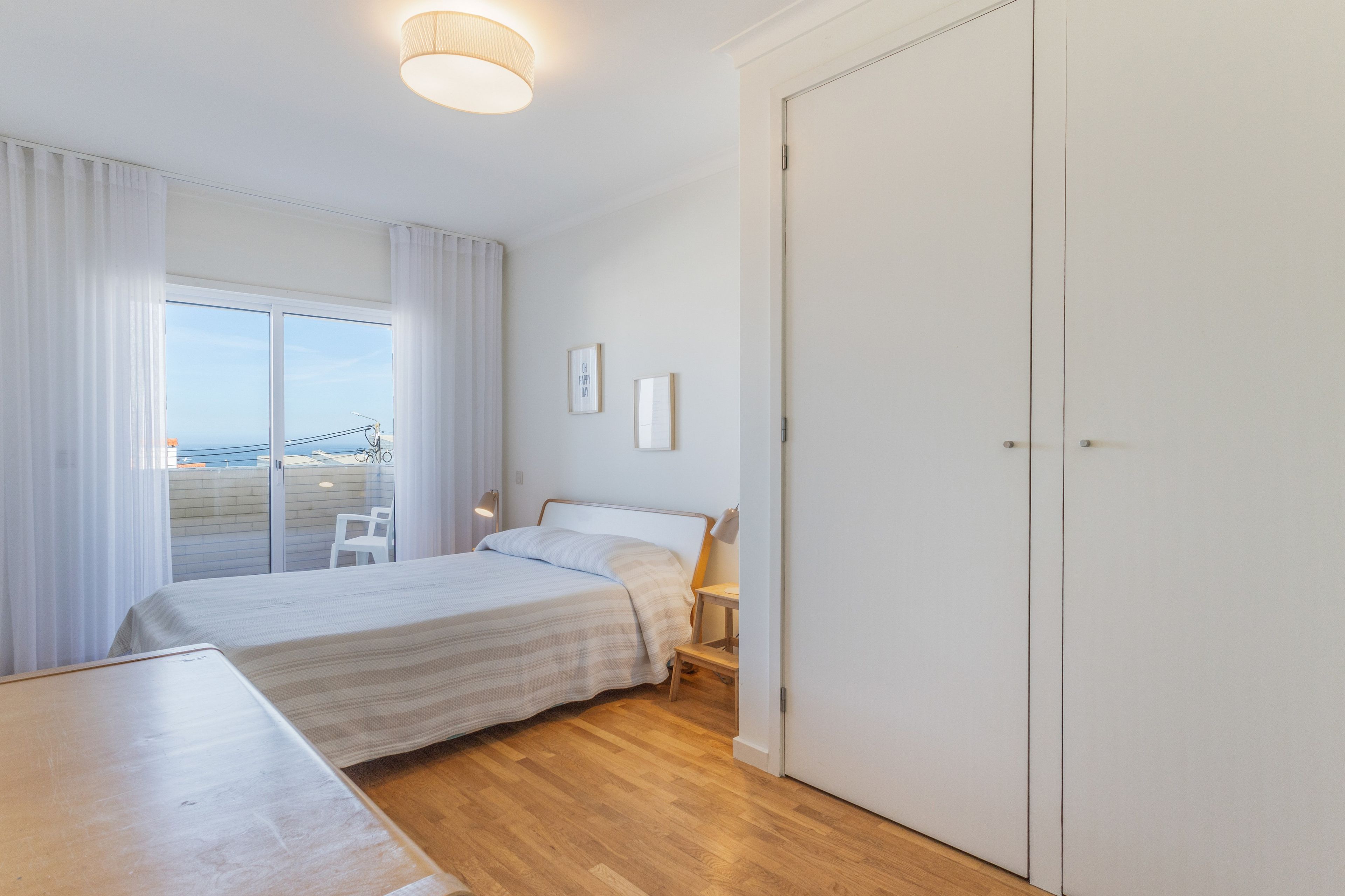A bedroom with a bed, a desk, a chair, a wardrobe, and a view through a window.