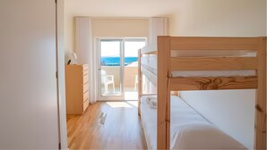 Una habitación con una cama litera, un aparador de madera, una puerta corrediza de vidrio que da a un balcón con vista al mar y una lámpara en el techo.