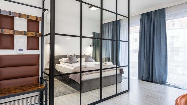 A modern hotel room with a large bed, a seating area with a bench and shelves, and a view through a glass partition to the outside.