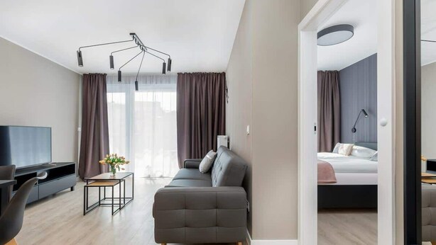 A modern living room with a sofa, a TV, and a small table. There is a bed in the adjacent room.