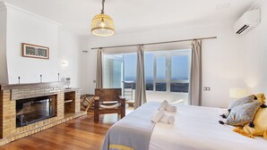 Ein Schlafzimmer mit Kamin, einem Bett mit Handtüchern, einem Sessel und Blick durch große Fenster.