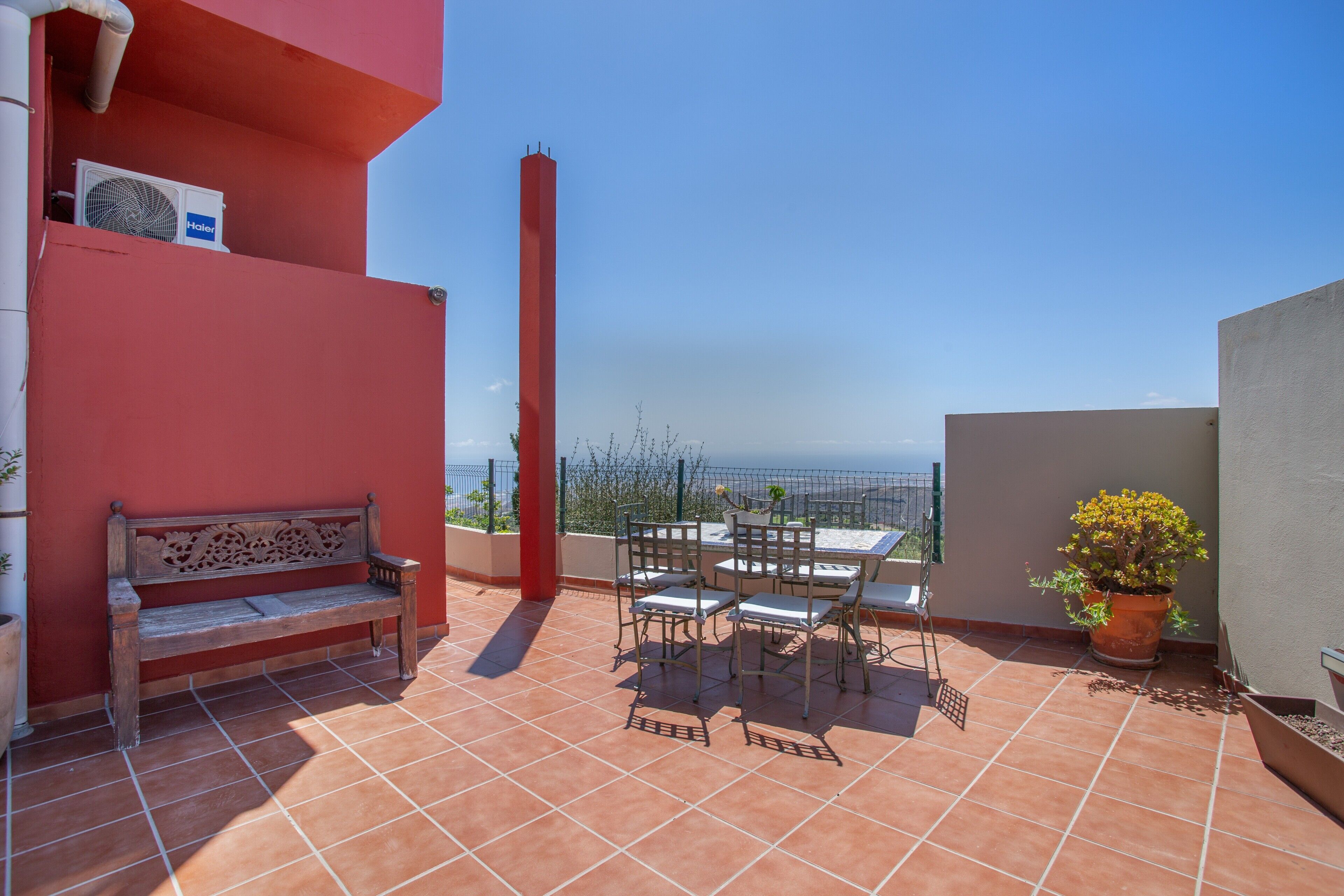 Una terraza con un muro rojo, un banco, una mesa con sillas y una planta en maceta.