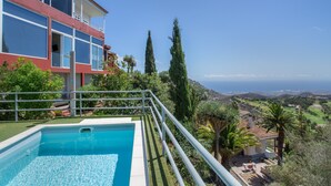 Una piscina amb vista a l'oceà, un edifici de dos pisos amb balcons, i un paisatge ple d'arbres i turons.