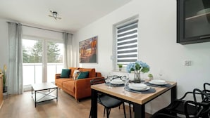 A modern living room with a wooden dining table set for two, a sofa with cushions, a large window with blinds, and a painting on the wall. - Stylish Apartment in Poznan | Terrace | Parking (Poznań)