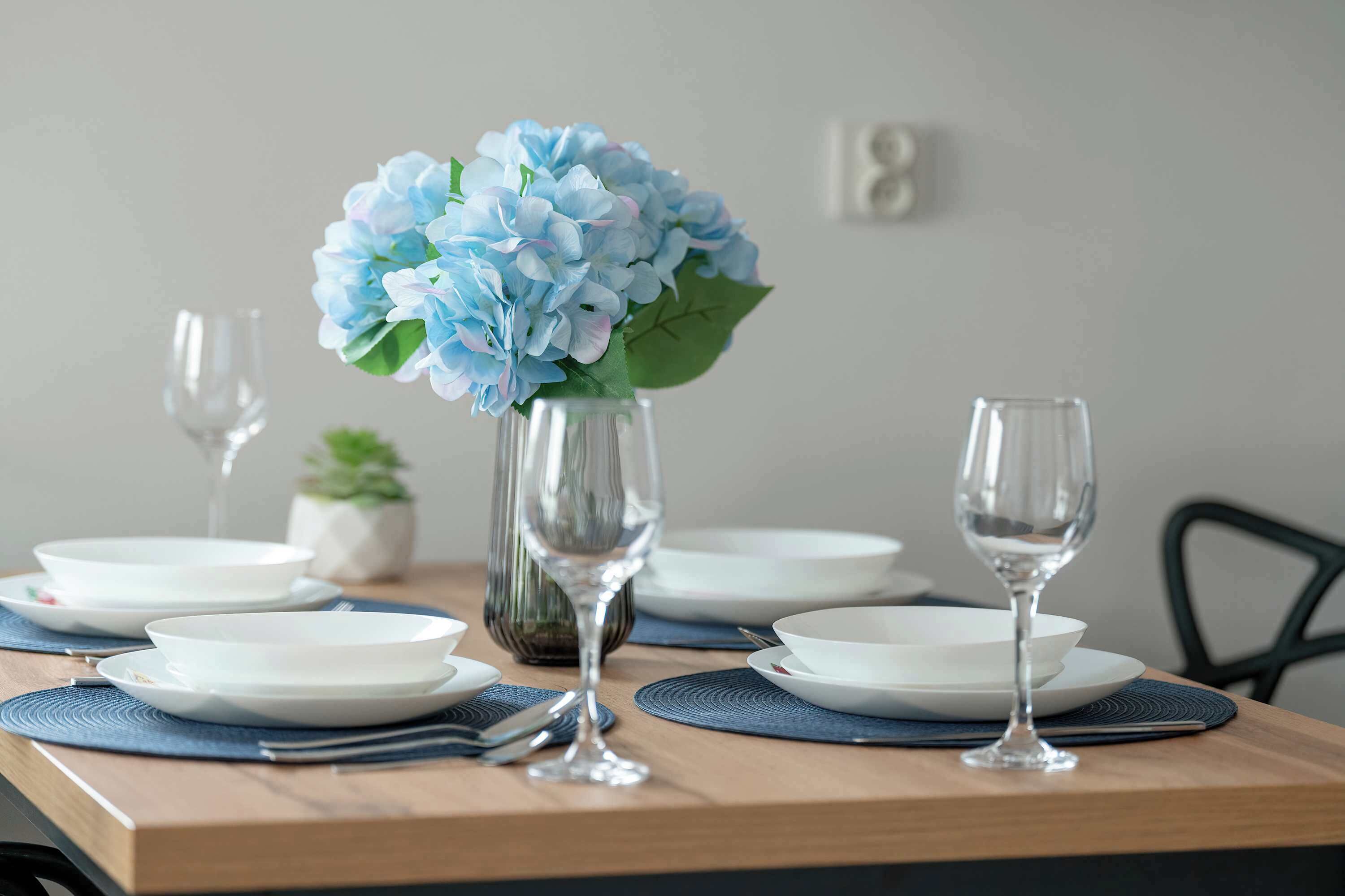 Een eettafel gedekt met witte borden, blauwe placemats en een vaas met blauwe bloemen.