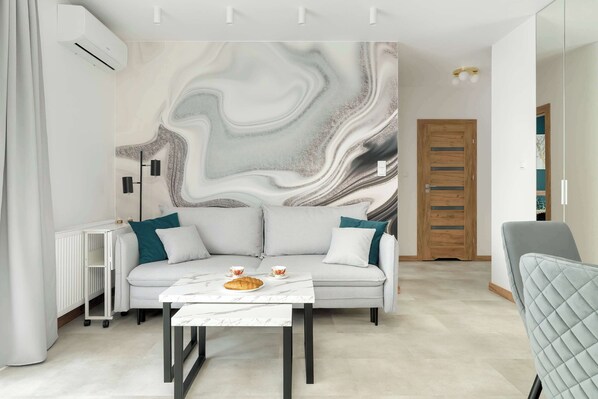 Ein modernes Wohnzimmer mit Wand im Marmoroptik, einem grauen Sofa mit türkisblauen Kissen, einem weißen Couchtisch und einem grauen Sessel.