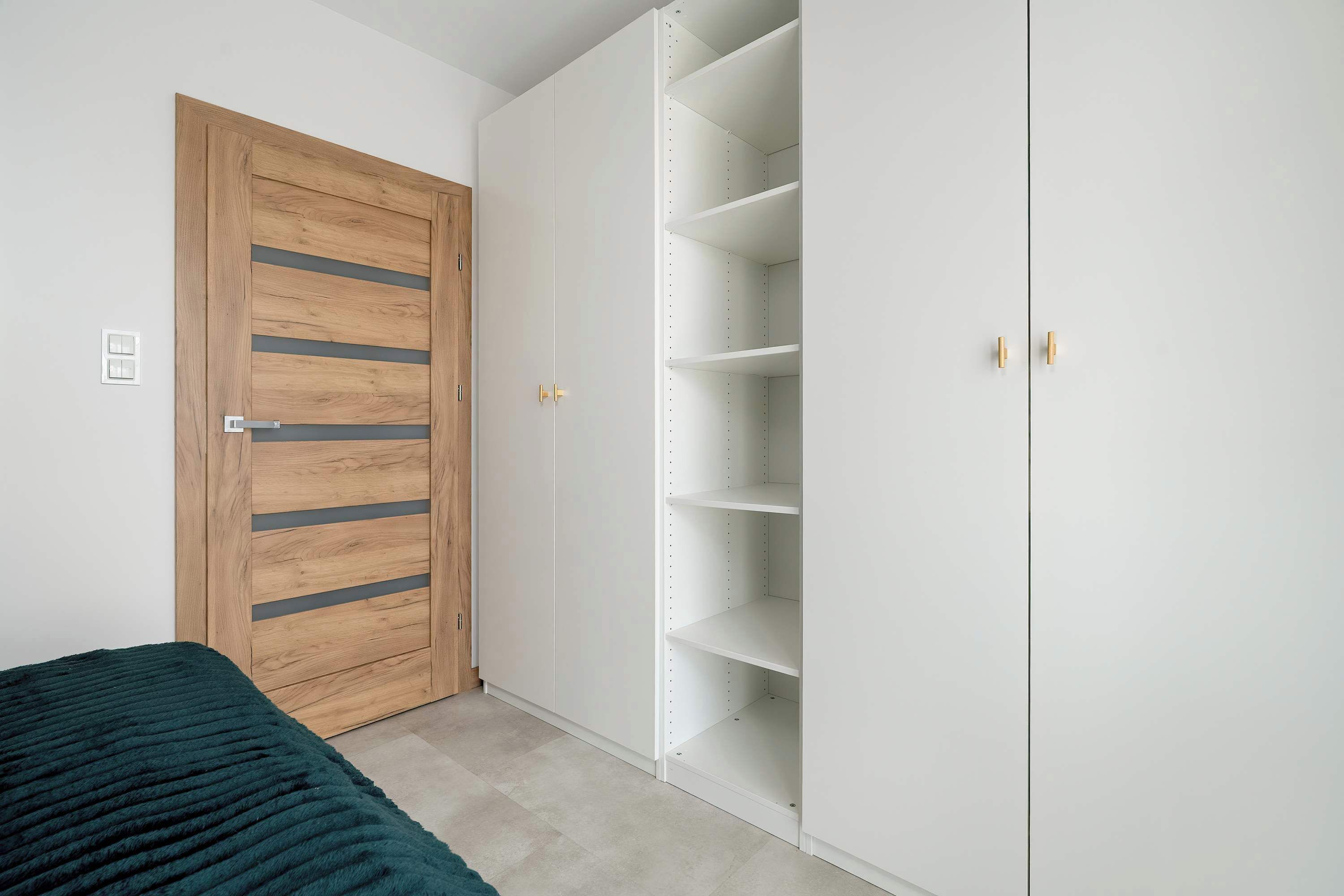 Ein Schlafzimmer mit einer Holztür, einem weißen Kleiderschrank mit vielen Ablagen und einer grünen Bettdecke.