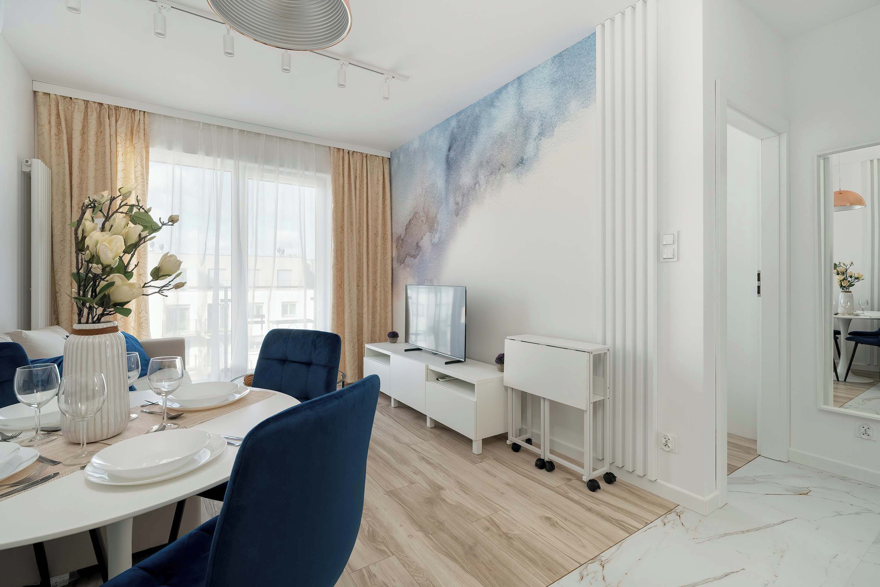 Un'area pranzo moderna con un tavolo bianco apparecchiato per quattro persone, sedie blu, un vaso con fiori bianchi e un grande quadro appeso al muro.