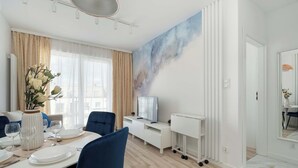 Un espace repas moderne avec une table blanche dressée pour quatre personnes, des chaises bleues, un vase avec des fleurs blanches et un grand tableau accroché au mur.