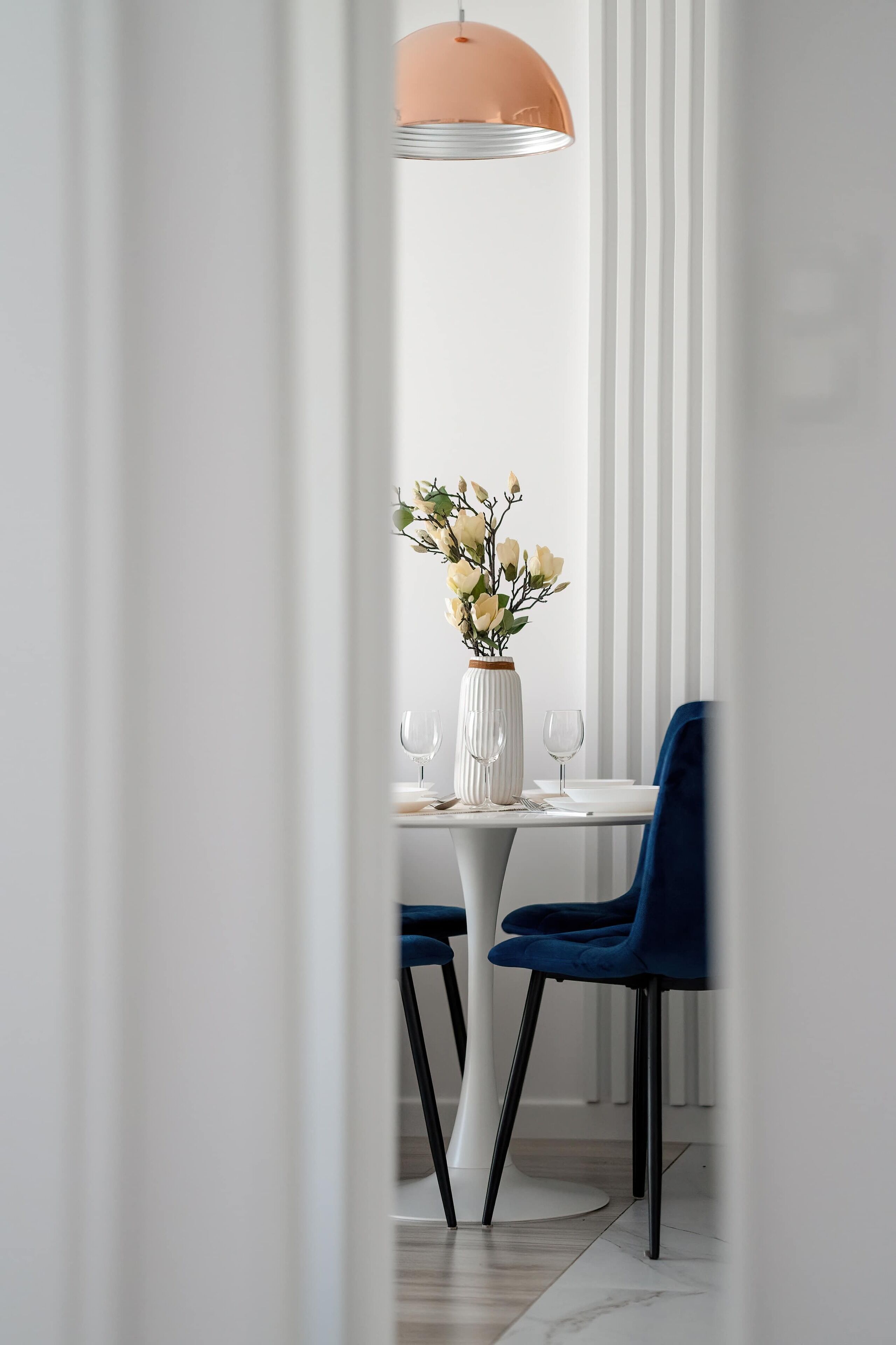 A dining area with a white table, two blue chairs, a white vase with flowers, and a pendant light.