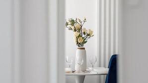 Un coin repas avec une table blanche, deux chaises bleues, un vase blanc orné de fleurs et un luminaire suspendu.