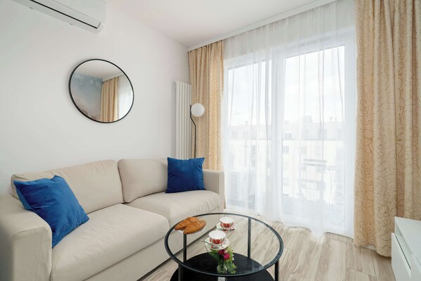 A modern living room with a beige sofa, blue cushions, a glass coffee table with a croissant, teacups, and a vase with flowers. - Stylish Apartment with Balcony | Parking | Close to Malta Lake (Poznań)