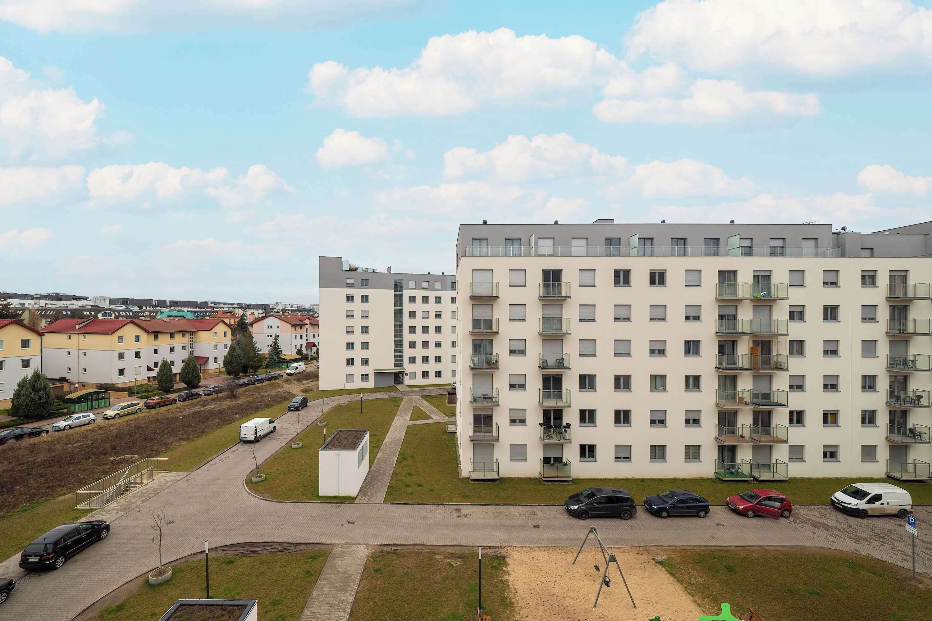 A multi-story residential building with balconies, a parking area with cars, and a clear sky.