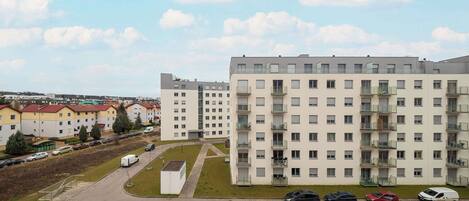 A multi-story residential building with balconies, a parking area with cars, and a clear sky.