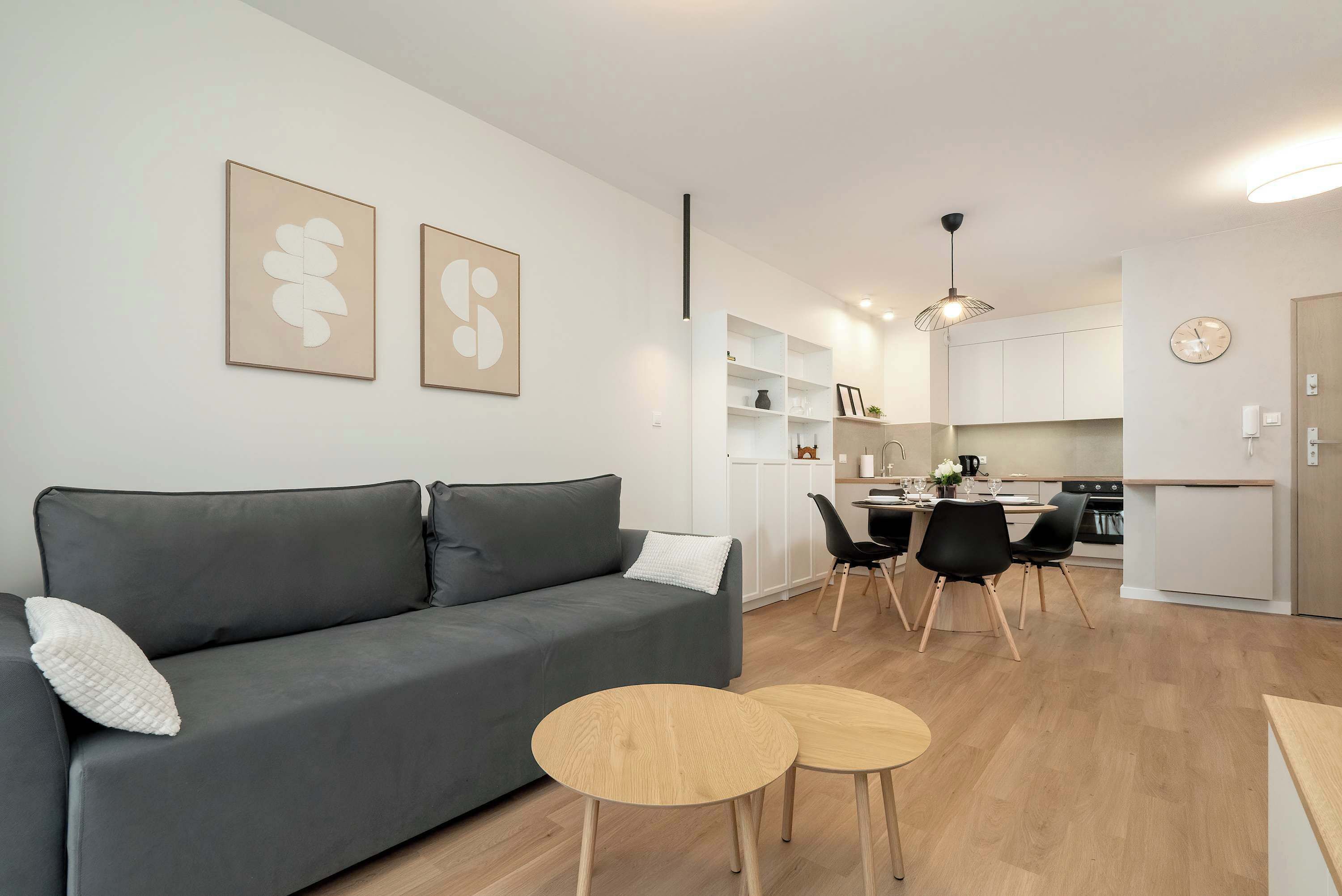 A modern living room with a grey sofa, two wooden side tables, and a dining area with a black chair and a white cabinet.