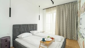 A neatly made bed with white linens, a grey headboard, and a wooden bedside table with a tray holding a cup, a plate, and a small plant. - Modern Apartment in Poznan | Balcony, Parking (Poznań)