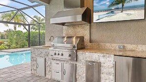 A modern outdoor kitchen with a stainless steel grill, built-in cabinets, and a large window overlooking a pool and tropical scenery.