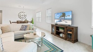 A modern living room with a white sofa, a glass coffee table, a wooden TV stand, and a large flat-screen TV displaying a beach scene.