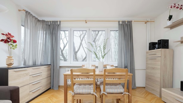 A modern dining area with a wooden table and chairs, a vase with flowers, and a view through large windows.