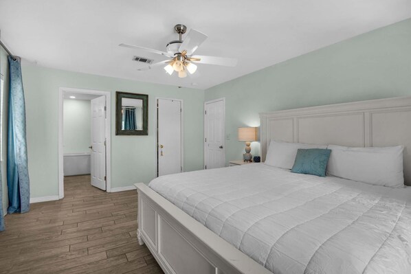 A bedroom with a bed, a ceiling fan, and a door leading to a bathroom. - Seaside Serenity (Pensacola Beach)