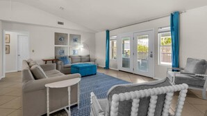 Un salon spacieux avec un grand canapé, un pouf bleu et un fauteuil blanc. On y trouve un ventilateur de plafond, une porte donnant sur l’extérieur et une fenêtre avec des rideaux bleus.