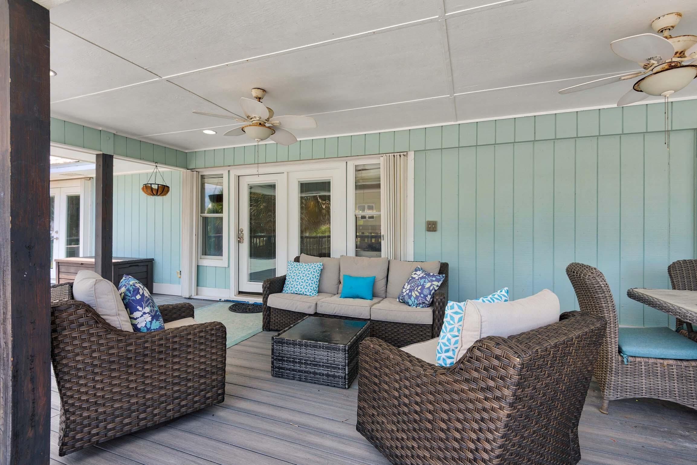 A covered outdoor seating area with wicker furniture, a glass-top coffee table, and ceiling fans.