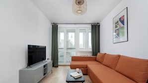 Ein modernes Wohnzimmer mit Flachbildfernseher, einer grauen Unterhaltungseinheit, einem orangefarbenen Sofa, einem kleinen Tisch mit Buch und Tasse sowie einem großen Fenster mit Vorhängen.