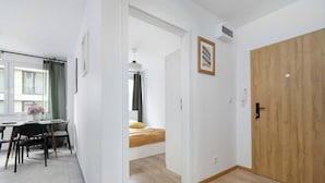 Ein modernes Zimmer mit Holztür, einem Bett mit gelber Decke, einer Essecke mit Tisch und Stühlen sowie einem Fenster mit Vorhängen.