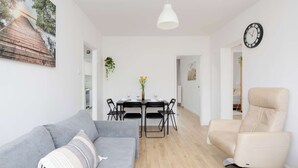 A modern living room with a grey sofa, a beige recliner, a dining table with chairs, and a wall-mounted clock.