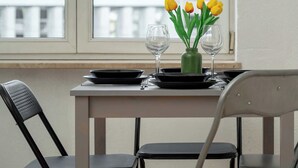 A dining table with a vase of yellow tulips, two wine glasses, and a green bottle. There are two chairs, one black and one grey, and a window with blinds.