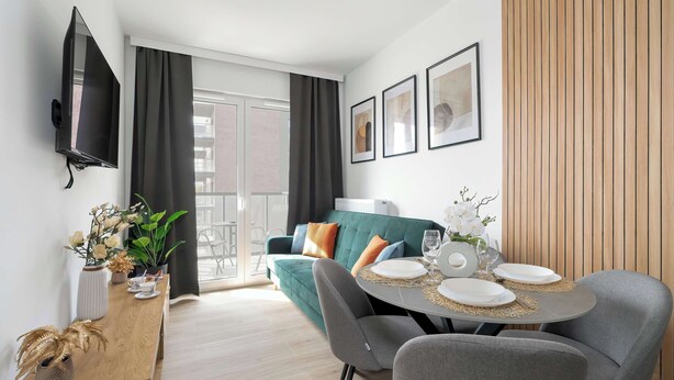 Ein modernes Wohnzimmer mit einem grünen Sofa, grauen Sesseln, einem Esstisch für zwei Personen, einem flachbildfernseher an der Wand und einem Balkon mit Blick.