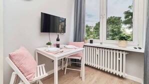 Ein kleines Zimmer mit einem weißen Tisch, einem Stuhl, einem an der Wand montierten Fernseher, einem Fenster mit Blick ins Grüne und einem Heizkörper unter dem Fenster.