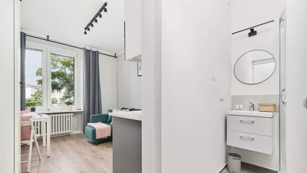 Ein modernes Zimmer mit Fenster, einer Couch, einem kleinen Tisch und einem Badezimmer mit Spiegel und Waschbecken.